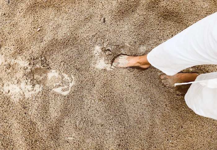 Aesthetic stock image shows a woman wearing white trousers walking on a sandy beach. The image is purely for decorative use only to support the blog post on how to become your best self so you can go on to live your best life