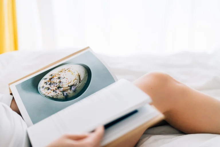 An aesthetic stock image showing an open recipe book on a lap of a woman who sits on a bed. The image is purely for decorative use only to support the blog post on the best nutrition books