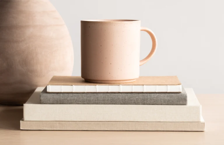 Aesthetic stock image that shows a stack of books and notebooks on a wooden table with a beige porcelain mug on the top. The image is purely for decorative use only to support the post on the best productivity books