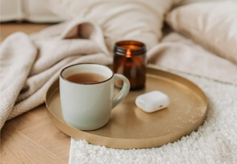 Aesthetic stock image showing a mug of tea, headphones and a candle on a gold tray on top of a blanket. The image is purely for decorative us only to support the blog post on feel-good burnout activities to do when you’re burnt out.