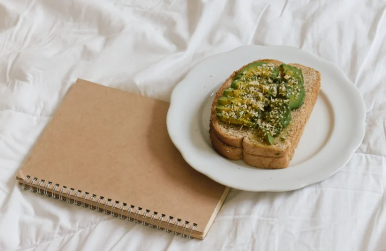 Aesthetic stock image shows avocado toast on a plate on a bed with white sheets next to a brown hardback notebook. The image is purely for decorative use only to support the blog post on Defining Wellness; The 6 Core Pillars of Health and Wellness and How To Practice Them