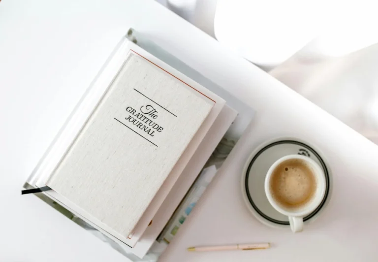 Aesthetic stock image shows a gratitude journal on a stack of notebooks on a white table top. The image is purely for decorative use only to support the blog post on gratitude journal prompts
