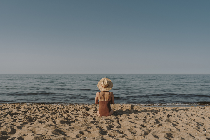 Aesthetic lifestyle images shows a woman wearing a brown swimsuit sitting on the beach with herself. The image is purely for decorative use only to support the blog post on how to let go of fear and travel solo in your thirties