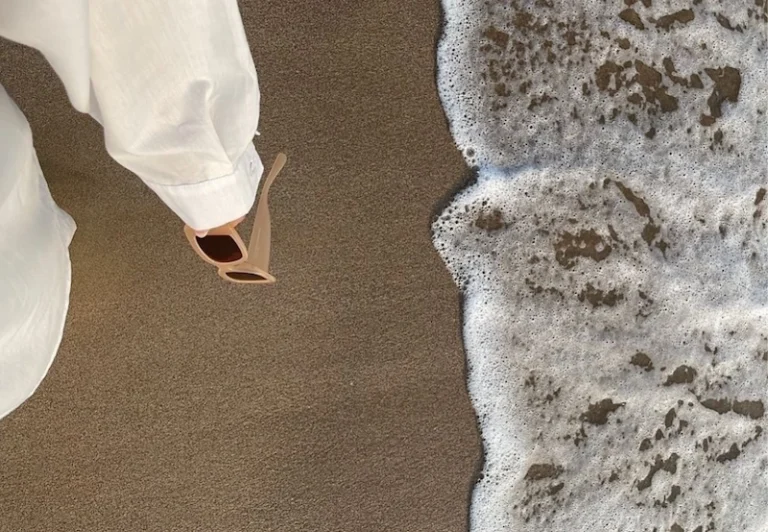 Aesthetic stock image showing an arm covered by a white shirt sleeve holding a pair of sunglasses walking along the beach. The image is purely for decorative use only to support the blog post on how to enjoy your life.