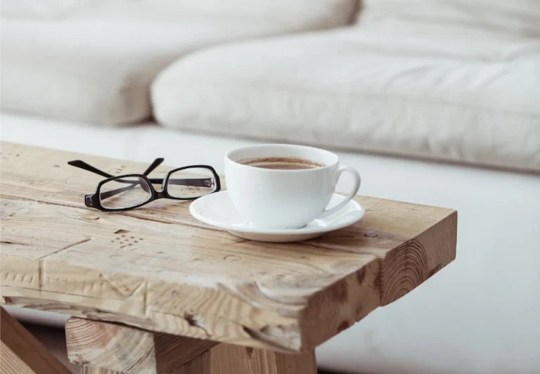 Aesthetic stock image showing a mug of coffee and a pair of glasses on top of a wooden bench in a living room. The image is purely for decorative use only to support the blog post on 101 habits to protect your energy and keep burnout away