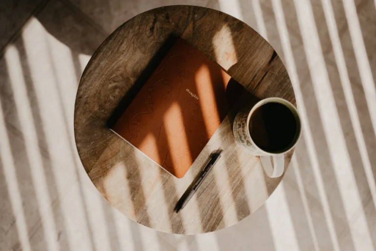 An aesthetic stock image showing a brown leather notebook on a wooden stool with a pen and a mug of coffee. Image is purely for decorative use only to support the blog post on the best reflective journals.