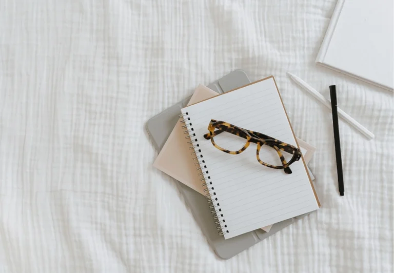 Aesthetic stock image showing a blank notebook on a bed with dark framed glasses lying on top. The image is purely for decorative us only to support the blog post on journal prompts to help with the question “should I quit my job?”