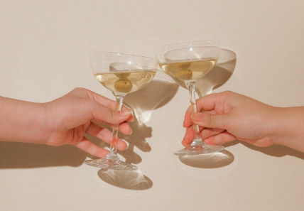 Aesthetic stock image showing two hands clinking a martini glass each with champagne against a plain white wall. The image is purely for decorative use only to support the blog post on social self-care