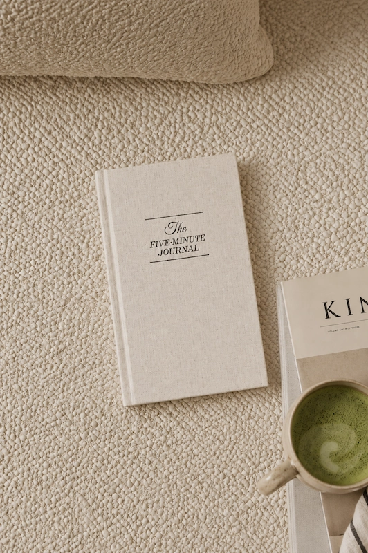 Aesthetic stock image showing The Five Minute Journal on a beige boucle carpet next to a mug of matcha latte.